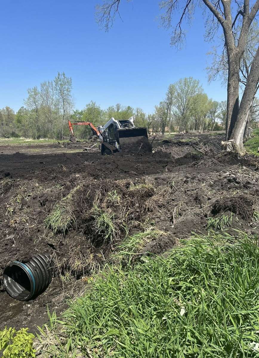 Skid steer and excavator grading dark soil beside a drainage culvert pipe